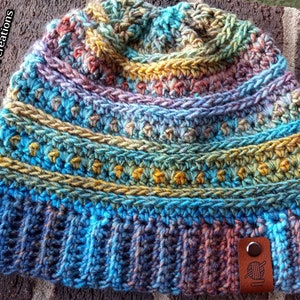 Pu&ograve; includere: Un berretto lavorato a uncinetto colorato con una toppa in pelle marrone che presenta un gomitolo di lana e un ago. Il berretto ha un design multicolore con sfumature di blu, verde, giallo e arancione.