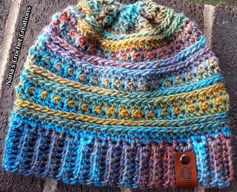 Pu&ograve; includere: Un berretto lavorato a uncinetto colorato con una toppa in pelle marrone che presenta un gomitolo di lana e un ago. Il berretto ha un design multicolore con sfumature di blu, verde, giallo e arancione.