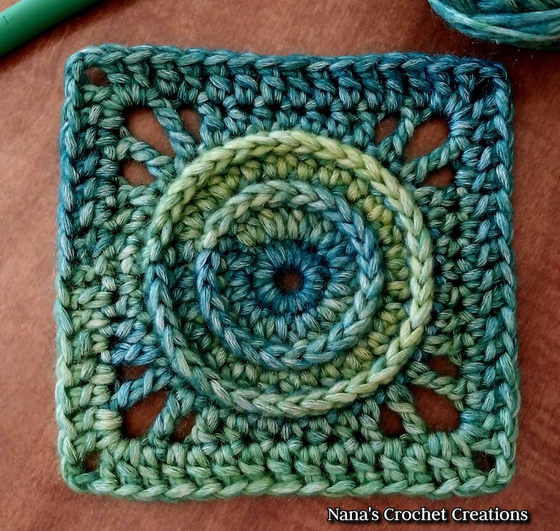 May include: A green, blue, and yellow crochet square with a circular design in the center. The square has a scalloped edge and is made with a variety of stitches.