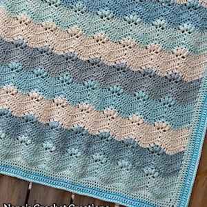 May include: A crocheted baby blanket with a wavy pattern in shades of blue, green, and beige. The blanket has a turquoise border. "Nana's Crochet Creations" is written in the bottom left corner.