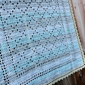 Op de afbeelding: Een gehaakt babydekentje in tinten blauw en groen met een witte gehaakte rand. Het dekentje is gemaakt met een textuur-steekpatroon en heeft een subtiel ombre-effect.