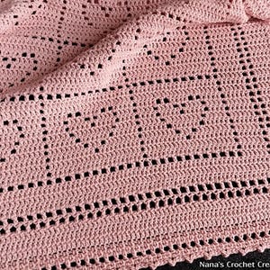 Puede incluir: Una manta de ganchillo rosa claro hecha a mano con un patrón de corazón. La manta presenta un diseño repetido de corazones abiertos y un borde decorativo. La textura es visible, mostrando la detallada artesanía. La manta es suave y acogedora.