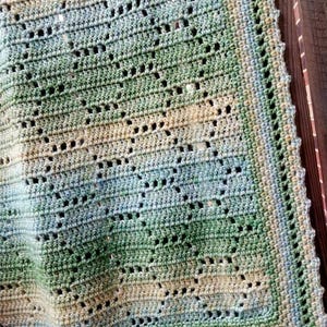 May include: A crochet blanket featuring a variegated yarn pattern in shades of green, blue, and cream. The textile displays a repeating geometric openwork design with a decorative border. The text Nana's Crochet Creations is visible.