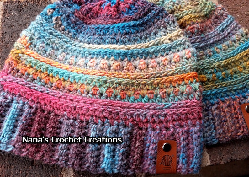 Pu&ograve; includere: Due cappelli all'uncinetto colorati con un'etichetta in pelle marrone che dice "Nana's Crochet Creations" e un'icona di filato.
