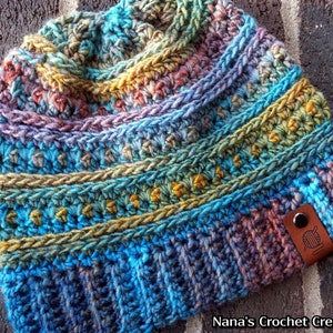 Pu&ograve; includere: Un berretto lavorato a maglia all'uncinetto colorato con un'etichetta in pelle marrone che dice "Nana's Crochet Creations".