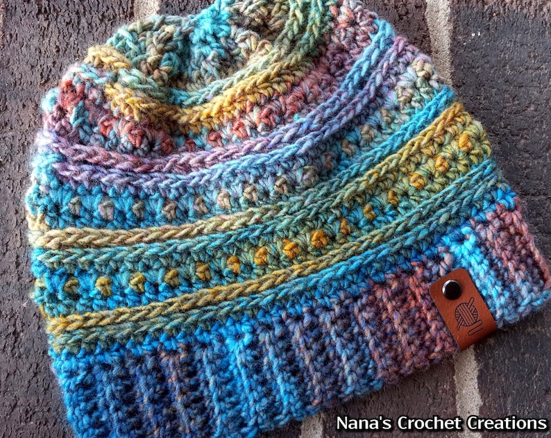 Pu&ograve; includere: Un berretto lavorato a maglia all'uncinetto colorato con un'etichetta in pelle marrone che dice "Nana's Crochet Creations".