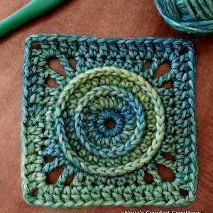 May include: A crocheted square with a circular design in the center. The square is made with a gradient yarn that transitions from green to blue. The center circle has three concentric rings, each with a different shade of green and blue.