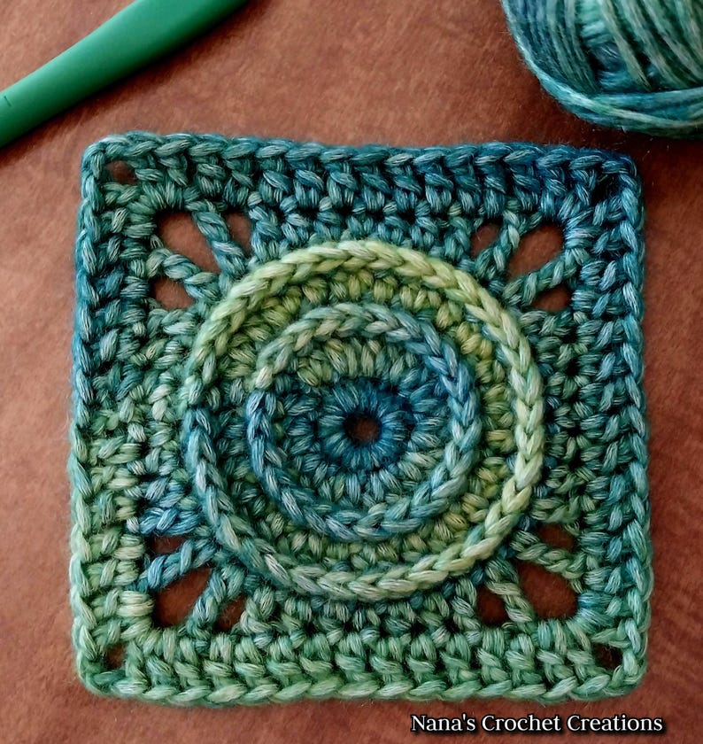 May include: A crocheted square with a circular design in the center. The square is made with a gradient yarn that transitions from green to blue. The center circle has three concentric rings, each with a different shade of green and blue.