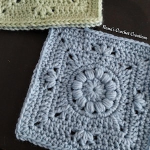 May include: Close-up of two crocheted squares. The bottom square is light blue with a central floral design and a textured border. The top square is a lighter green. The text "Nana's Crochet Creations" is visible.