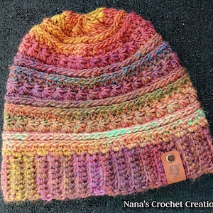 May include: A crocheted beanie hat with a multi-colored yarn in shades of orange, red, pink, green, and brown. The hat has a textured stitch pattern and a leather tag with a crochet hook and yarn symbol.