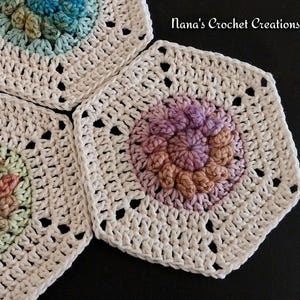 May include: Close-up of three handmade crochet coasters. Each coaster features a white, openwork design with a central floral motif in shades of blue, pink, and peach. The text "Nana's Crochet Creations" is visible in the upper right corner.