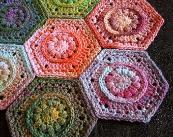 May include: A close-up of a crocheted hexagon blanket with a variety of colors, including green, pink, blue, and purple. The blanket is made up of individual hexagons that are joined together. The hexagons have a raised, textured design in the center.