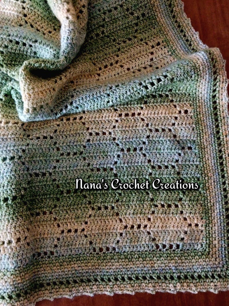 May include: A handmade crochet blanket featuring a variegated color palette of sage green, forest green, and cream. The textile displays an openwork geometric pattern with a decorative border. The text Nana's Crochet Creations is visible.