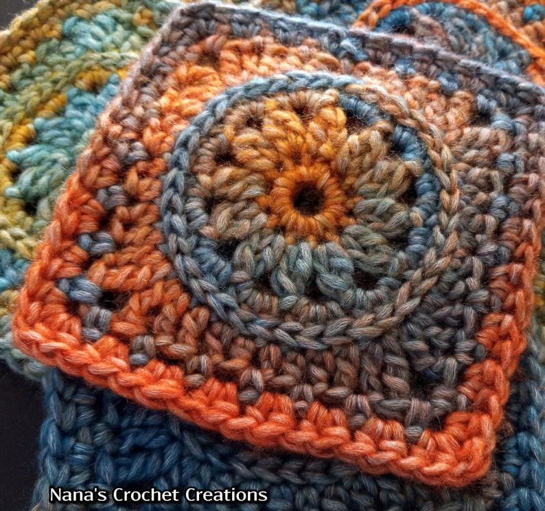May include: A close-up of a crocheted square with a multi-colored design. The square is made with a variety of shades of blue, orange, and brown yarn. The center of the square features a circular design with a darker blue center.