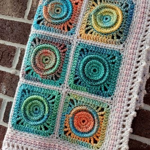 May include: A crocheted blanket with a multi-colored design featuring circular patterns. The blanket has a light pink border with a scalloped edge. The text "Nana's Crochet Creations" is visible in the top right corner of the image.