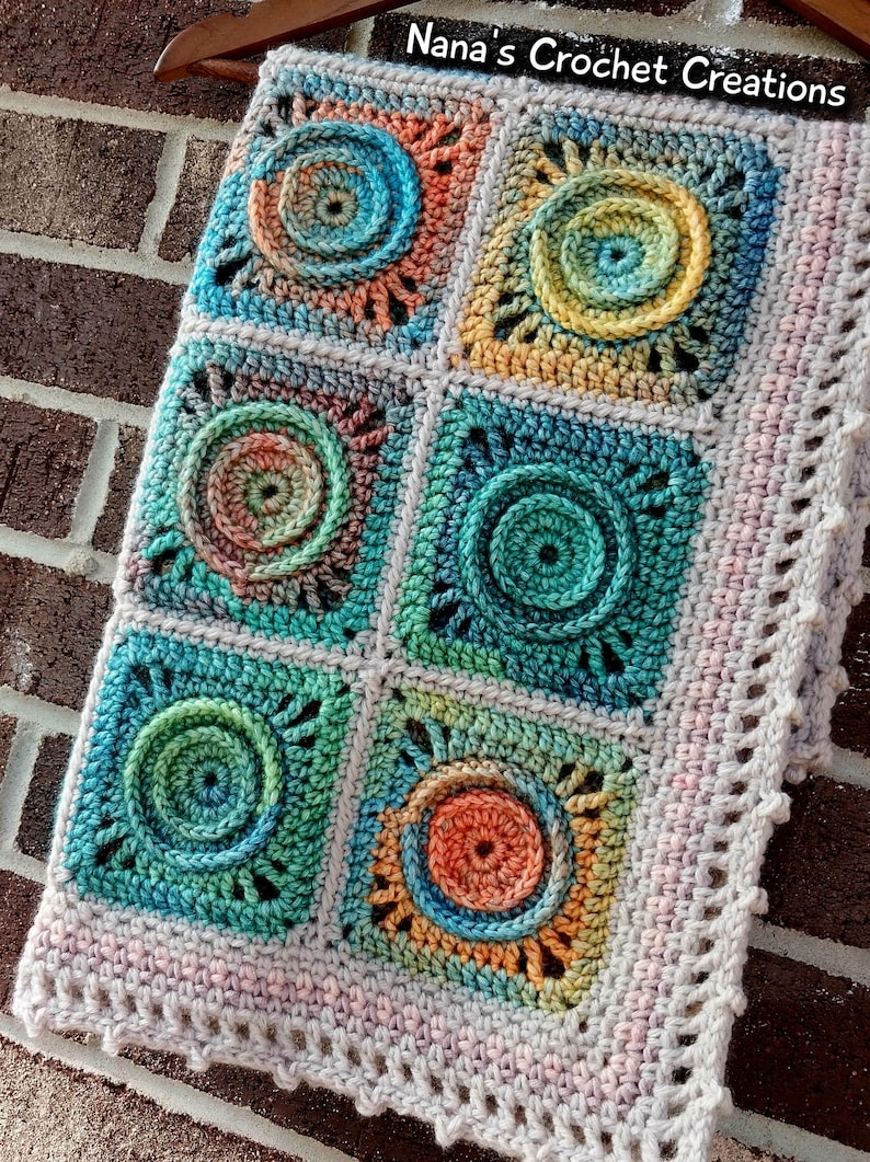 May include: A crocheted blanket with a multi-colored design featuring circular patterns. The blanket has a light pink border with a scalloped edge. The text "Nana's Crochet Creations" is visible in the top right corner of the image.
