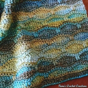 Op de afbeelding: Een close-up van een handgemaakte gehaakte deken met een golfachtig patroon. De deken heeft een mix van blauwe, groene, bruine en gele garens. De tekst "Nana's Crochet Creations" is zichtbaar in de rechter benedenhoek.