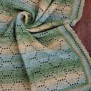 May include: A handmade crochet blanket featuring a gradient of green, cream, and light blue yarn. The textile displays an intricate openwork pattern with repeating geometric shapes and a scalloped border. The text Nana's Crochet Creations is visible.