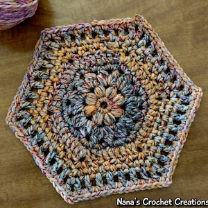 Może przedstawiać: Podkładka pod kubek w kształcie sześciokąta, wykonana na szydełku z wielokolorowej przędzy w odcieniach niebieskiego, pomarańczowego i różowego. Podkładka ma centralny wzór kwiatowy.