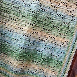 May include: A handmade crochet blanket featuring a repeating geometric pattern in a variegated color palette of soft green, pale blue, and cream. The textile has an openwork design with a finished border and the text Nana's Crochet Creations.