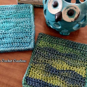 May include: Two handmade crochet coasters in shades of blue and green. One coaster is a solid blue with a wave pattern, and the other is a mix of green and blue. A turquoise owl-shaped candle holder is in the background. The text "Nana's Crochet Creations" is visible.