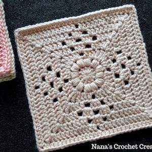 May include: A crocheted square with a delicate floral design in a light pink color. The square is made with a fine yarn and has a textured, lacy appearance. "Nana's Crochet Creations" is written in the bottom right corner.