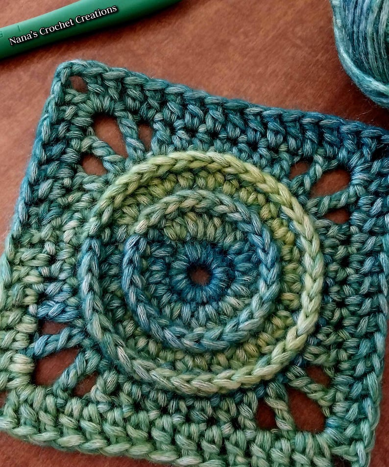 May include: A green and blue crochet square with a circular design in the center. The square has a scalloped edge and is made with a textured stitch.
