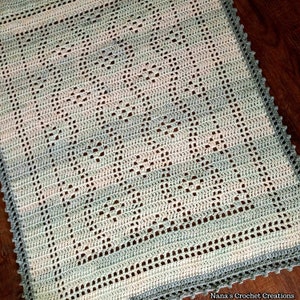 May include: A crocheted baby blanket in shades of green and white with a delicate lace border. The blanket features a repeating diamond pattern.