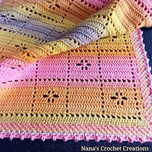 Pode incluir: Um cobertor de bebé em crochet multicolorido com um padrão rendado e uma borda rosa. O cobertor é feito com uma variedade de tons de rosa, laranja, amarelo e roxo.