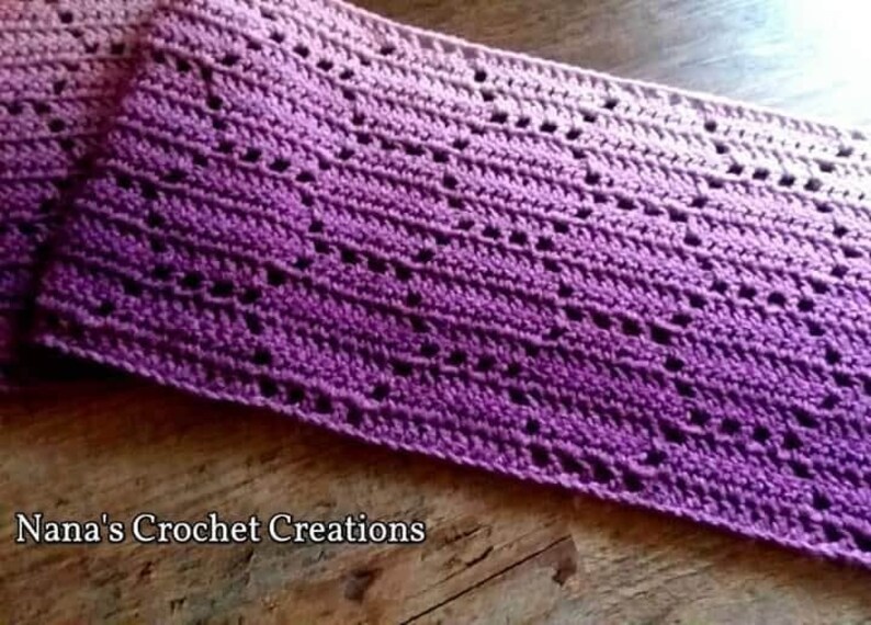 May include: A rectangular crochet scarf featuring an ombre gradient that transitions from light pink to deep purple. The fabric displays a repeating openwork geometric pattern throughout. The text Nana's Crochet Creations is printed in white.