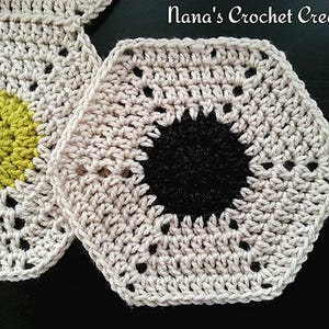 Puede incluir: Dos hexágonos de ganchillo, uno con un centro verde y el otro con un centro negro. Los hexágonos están hechos con hilo beige y tienen un patrón de encaje.