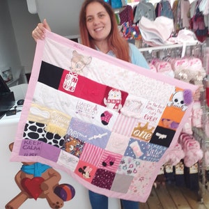 May include: A pink baby blanket with a variety of colorful patches sewn together. The patches feature different designs, including animals, words, and numbers. The blanket is being held by a person in a blue shirt.
