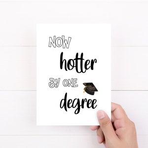 May include: A white graduation card with the text "Now hotter by one degree" in black script and a small graduation cap illustration. The card is held by a hand against a white wooden background.