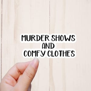 K&ouml;nnte beinhalten: Wei&szlig;er Aufkleber mit schwarzem Text, der "Murder Shows and Comfy Clothes" lautet.