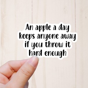 May include: White sticker with black text that reads "An apple a day keeps anyone away if you throw it hard enough."