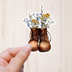 May include: A sticker featuring a watercolor illustration of brown boots filled with wildflowers. The boots have laces and are filled with white, yellow, and blue flowers. The sticker is held by a hand.