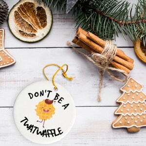 May include: A white round Christmas ornament with the text "Don't be a twatwaffle" and a cartoon waffle with a face and legs. The ornament is surrounded by gingerbread cookies and cinnamon sticks.
