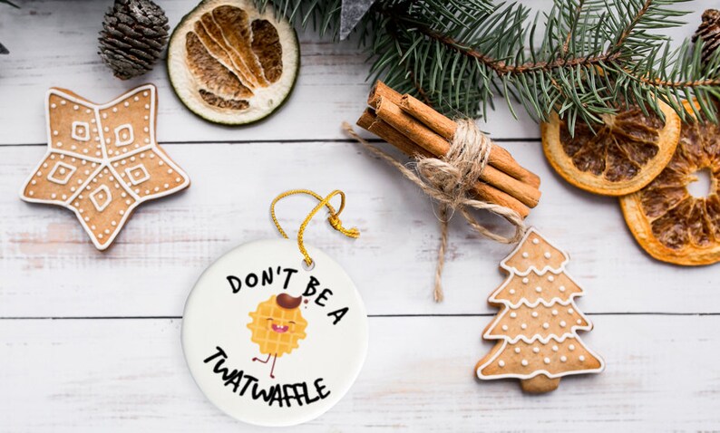 May include: A white round Christmas ornament with the text "Don't be a twatwaffle" and a cartoon waffle with a face and legs. The ornament is surrounded by gingerbread cookies and cinnamon sticks.