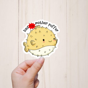 Puede incluir: Una pegatina blanca con un pez globo de dibujos animados con un cuerpo amarillo y manchas blancas. El texto "bad mother puffer" est&aacute; escrito en negro alrededor del pez. Una estrella roja con la palabra "oops" est&aacute; encima del pez.