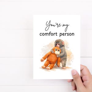 May include: A greeting card with the text "You're my comfort person" features a watercolor illustration of a gray monkey hugging an orange stuffed monkey. The card is held by a hand against a white background.