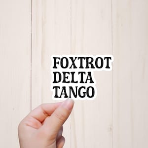 May include: White sticker with black text that reads "FOXTROT DELTA TANGO" on a wood background.