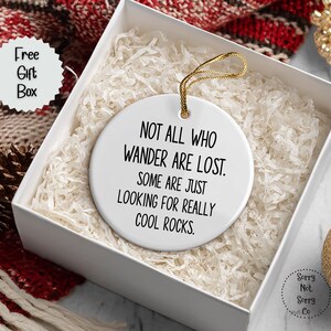 Puede incluir: Adorno de cer&aacute;mica blanca con cuerda dorada, dentro de una caja de regalo. El adorno tiene el texto "NOT ALL WHO WANDER ARE LOST. SOME ARE JUST LOOKING FOR REALLY COOL ROCKS." La caja est&aacute; llena de papel blanco y est&aacute; junto a una bufanda roja y blanca.