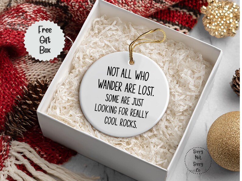 Puede incluir: Adorno de cer&aacute;mica blanca con cuerda dorada, dentro de una caja de regalo. El adorno tiene el texto "NOT ALL WHO WANDER ARE LOST. SOME ARE JUST LOOKING FOR REALLY COOL ROCKS." La caja est&aacute; llena de papel blanco y est&aacute; junto a una bufanda roja y blanca.