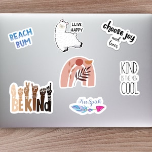 K&ouml;nnte beinhalten: Ein Laptop-Computer mit mehreren Aufklebern. Die Aufkleber zeigen verschiedene Designs und S&auml;tze, darunter ein Lama, ein Regenbogen, eine Feder und die Worte "Beach Bum", "Live Happy", "Choose Joy and Tacos", "Be Kind", "Kind is the New Cool" und "Free Spirit".