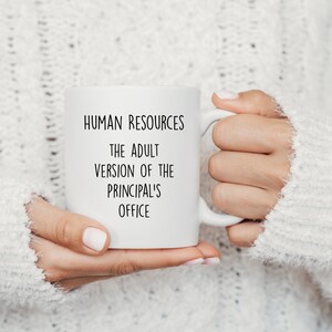 May include: White ceramic mug with the text "HUMAN RESOURCES THE ADULT VERSION OF THE PRINCIPAL'S OFFICE" in black lettering. The mug is held by a person wearing a white sweater.