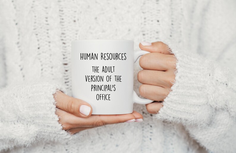May include: White ceramic mug with the text "HUMAN RESOURCES THE ADULT VERSION OF THE PRINCIPAL'S OFFICE" in black lettering. The mug is held by a person wearing a white sweater.