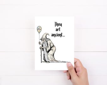 May include: A white greeting card featuring a whimsical illustration of an old wizard holding a balloon that says "Happy Birthday." The card reads "Thou art ancient..." in black text. The card is held by a hand.
