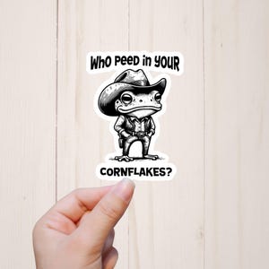 May include: Black and white sticker featuring a cartoon frog dressed as a cowboy. The frog wears a cowboy hat, shirt, and holster. Text above reads "Who peed in your" and below "Cornflakes?"