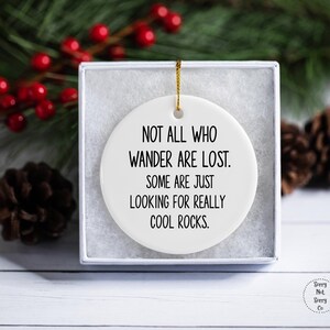 Puede incluir: Un adorno de cer&aacute;mica blanca con el texto "NOT ALL WHO WANDER ARE LOST. SOME ARE JUST LOOKING FOR REALLY COOL ROCKS." en negro. El adorno est&aacute; en una caja blanca, con una cuerda dorada para colgar. Pi&ntilde;as y bayas rojas en el fondo.