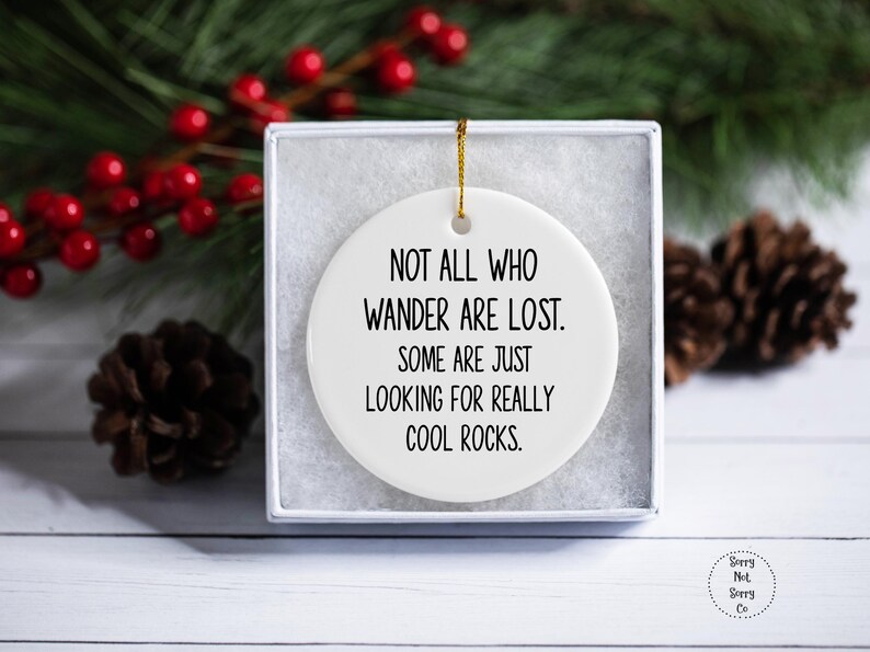 Puede incluir: Un adorno de cer&aacute;mica blanca con el texto "NOT ALL WHO WANDER ARE LOST. SOME ARE JUST LOOKING FOR REALLY COOL ROCKS." en negro. El adorno est&aacute; en una caja blanca, con una cuerda dorada para colgar. Pi&ntilde;as y bayas rojas en el fondo.